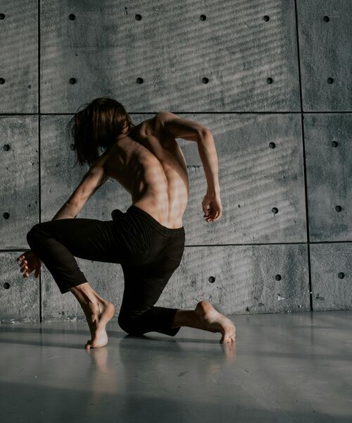 Eine Person mit nacktem Oberkörper bewegt sich tänzerisch auf dem Boden vor einer grauen Betonwand, das Licht wirft Schattenmuster auf ihren Körper.