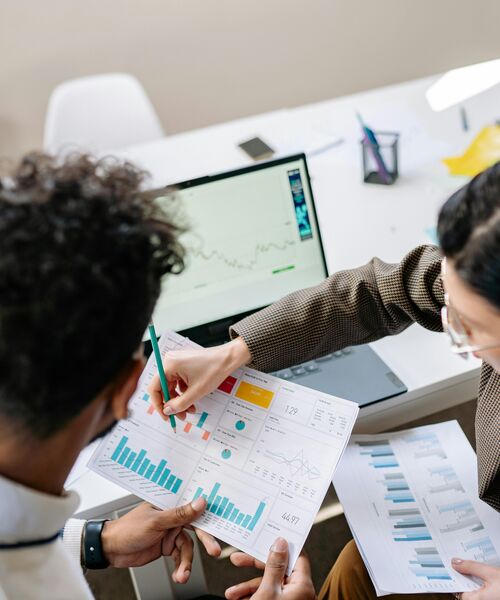 Zwei Personen analysieren gemeinsam Diagramme und Statistiken, während im Hintergrund ein Laptop mit Datenanzeige steht.