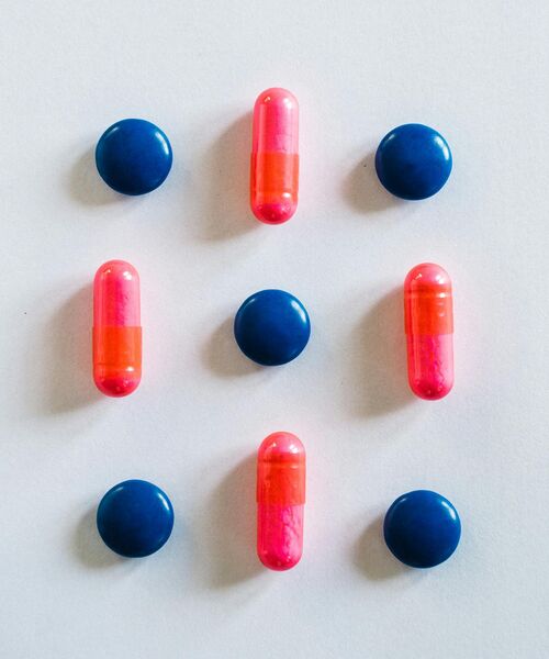 Mehrere blaue Tabletten und pink-orange Kapseln sind in einem symmetrischen Muster auf hellem Hintergrund angeordnet.