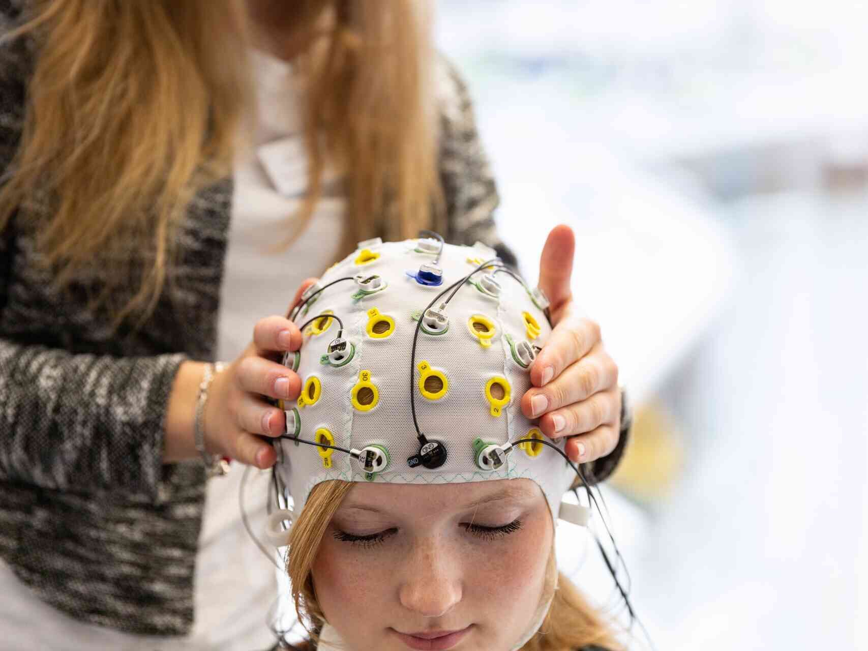 Eine junge Person trägt eine EEG-Messhaube mit Elektroden, während jemand die Sensoren überprüft.