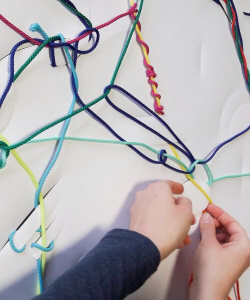 Zwei Hände knüpfen bunte Fäden an einer weißen Fläche zu einem Netz aus verschiedenen, miteinander verbundenen Schnüren.
