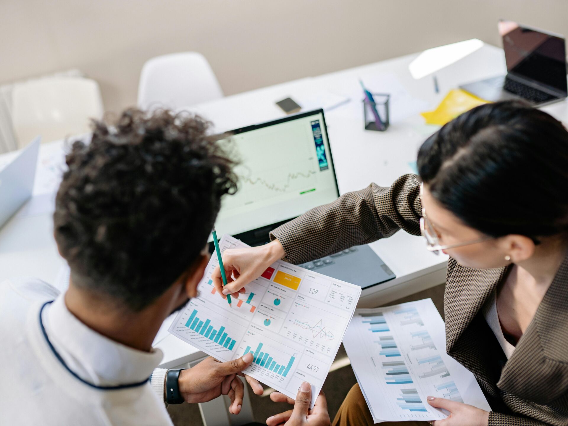 Zwei Personen analysieren gemeinsam Diagramme und Statistiken, während im Hintergrund ein Laptop mit Datenanzeige steht.