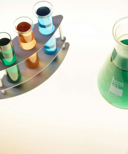 Mehrere Reagenzgläser mit farbigen Flüssigkeiten stehen neben einem Erlenmeyerkolben auf einem Labortisch.