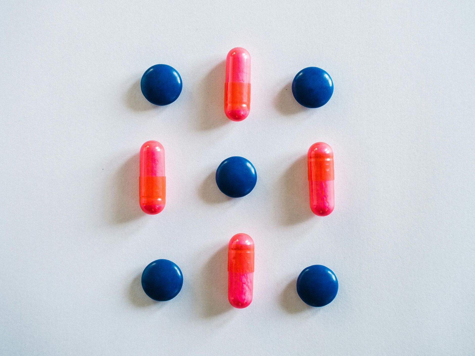 Mehrere blaue Tabletten und pink-orange Kapseln sind in einem symmetrischen Muster auf hellem Hintergrund angeordnet.