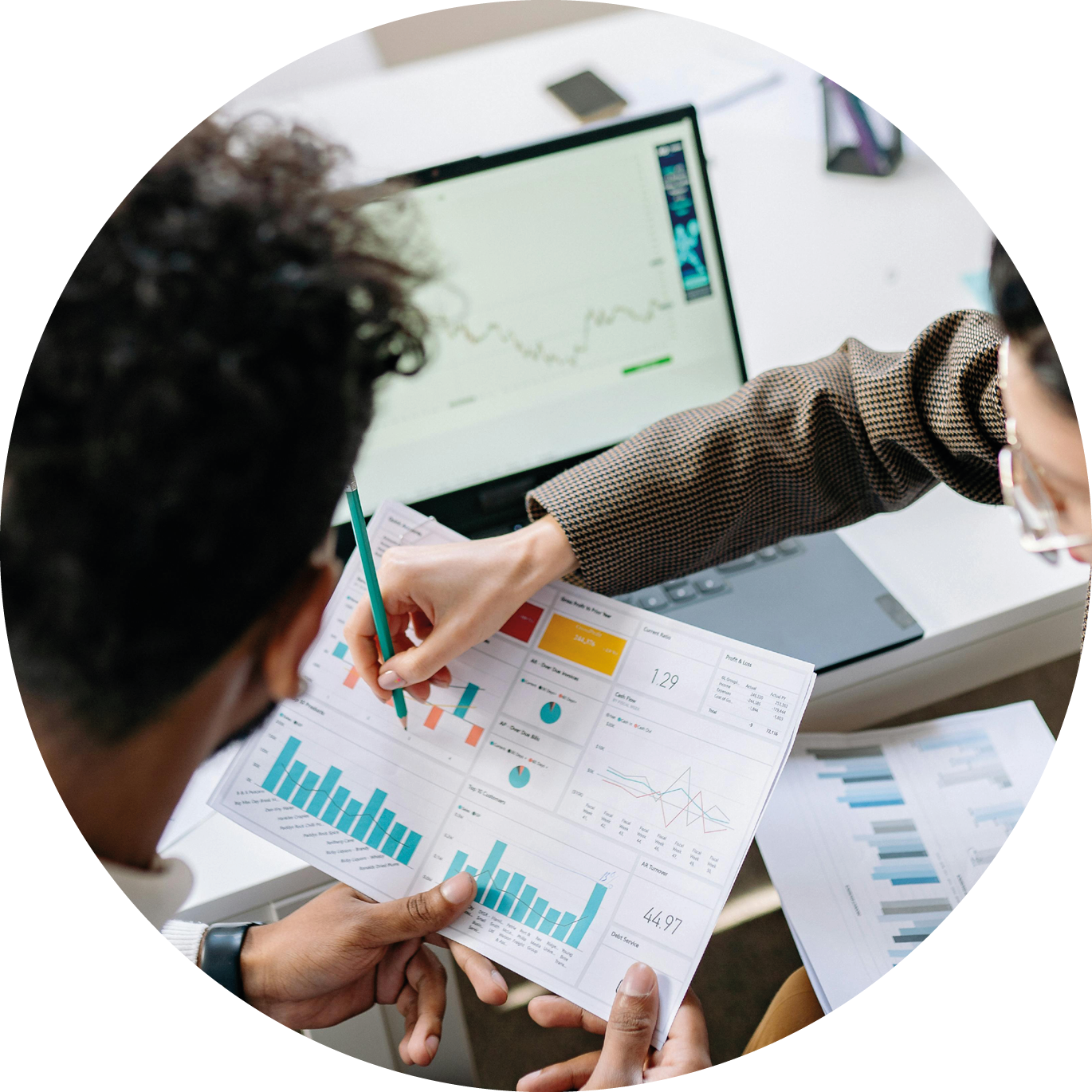 Zwei Personen besprechen Diagramme und Statistiken, während im Hintergrund ein Laptop mit Datenanalyse geöffnet ist.